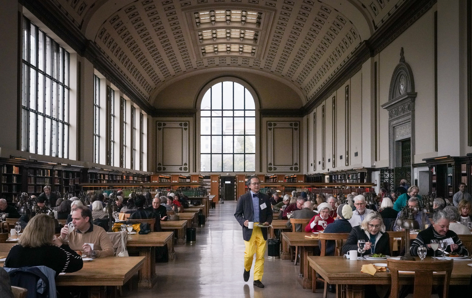 a man in yellow pants walks into the a crowded library with a plate of food