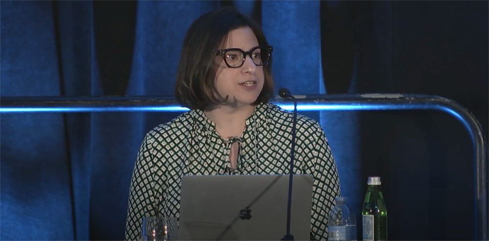 A woman with shoulder-length brown hair and black-rimmed glasses speaks at a podium during a conference. She is wearing a green and white patterned blouse with a keyhole neckline. A laptop is open in front of her, and a microphone is positioned nearby. The background features dark blue curtains, and the table holds a glass of water, a plastic water bottle, and a green glass bottle.
