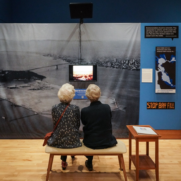 two guests sit on a bench listening in front of an exhibit