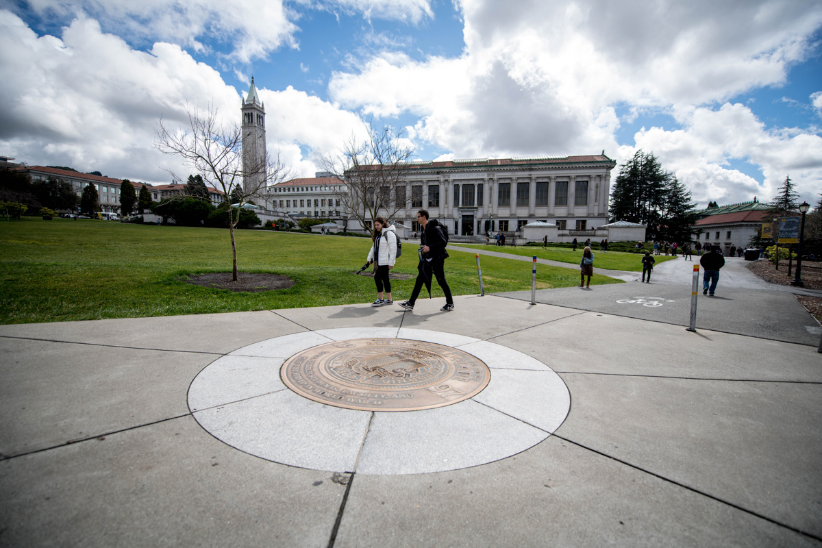 people walk on campus near a seal in the sidewalk with library in the background